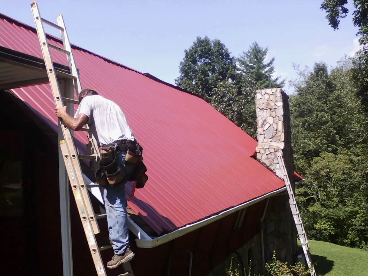 Roofer on a ladder beside a red metal roof and stone chimney — Soffit & Fascia Repair by Roofing Brothers in Westtown